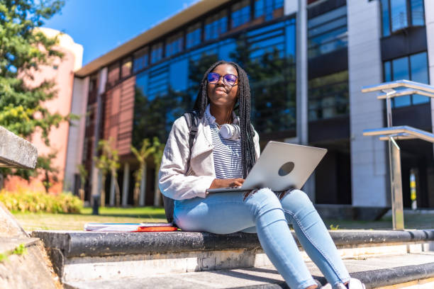 Distracted african woman looking up while using laptop sitting outside the university campus